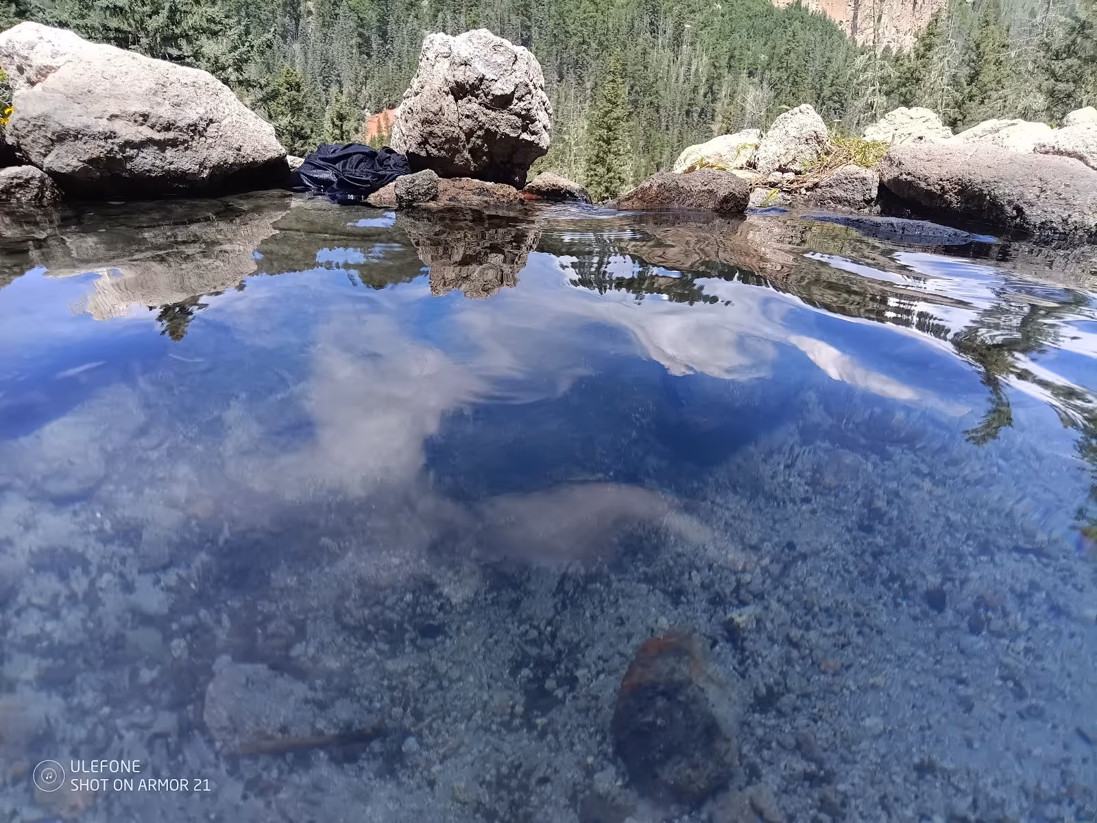 Clear water in a small hot spring pool reflects blue sky and surrounding evergreen trees with rocks forming the pool edge at San Antonio Ho.