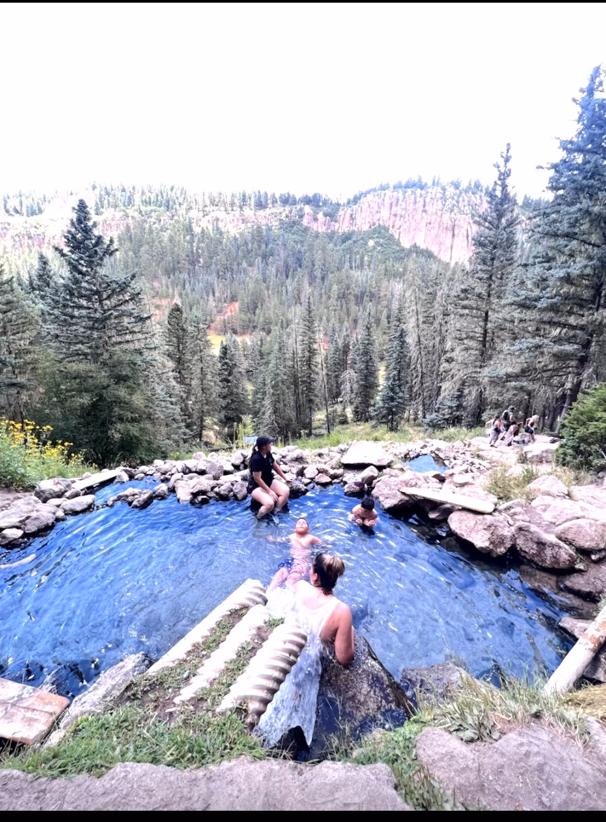 Group of people sit and soak in a rock-bordered hot spring pool high on a hillside with dense forest and red rock cliffs near Jemez Springs.