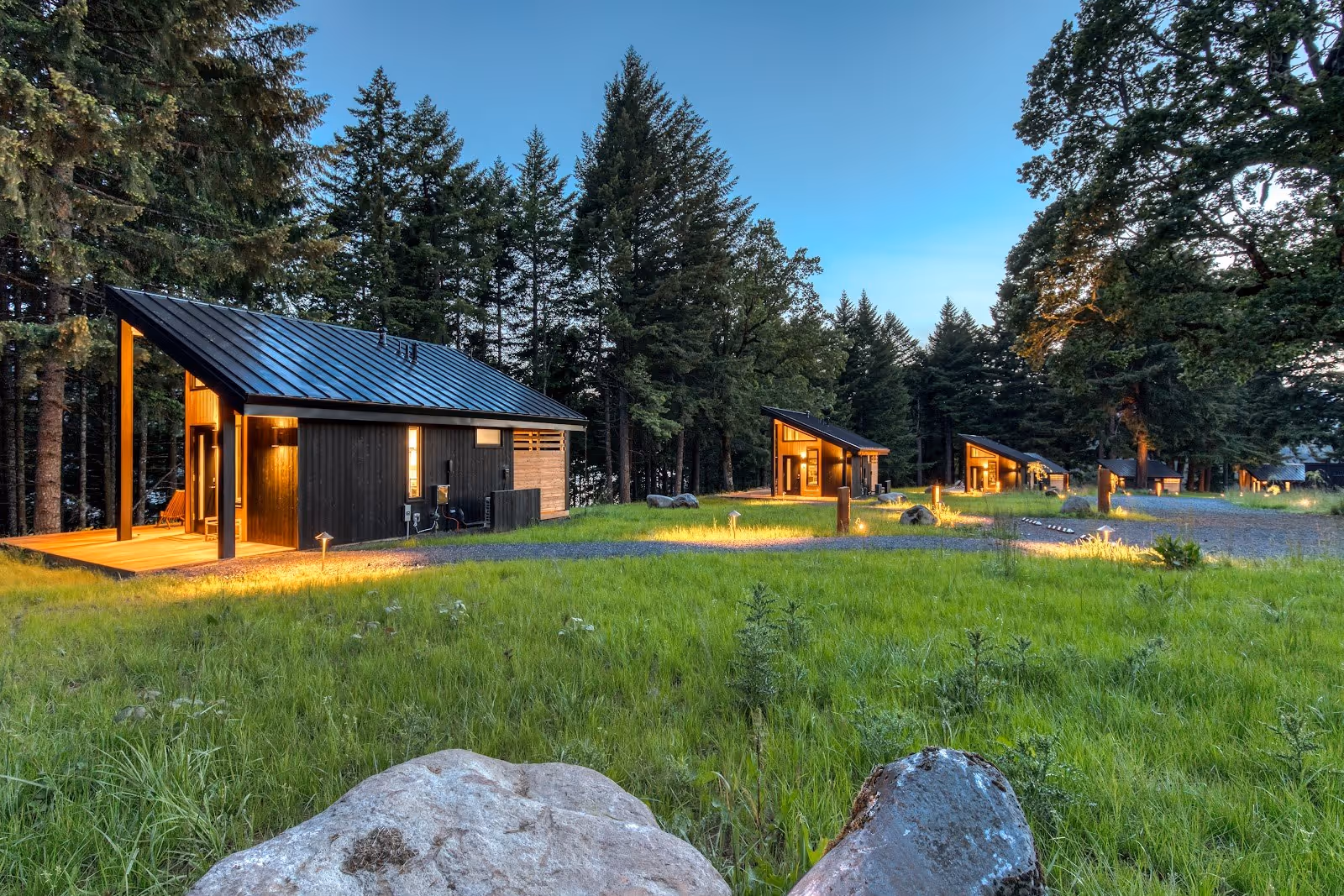 Exterior view of multiple modern dark wood cabins with metal roofs lit at dusk surrounded by grassy field and tall trees at Tenzen Springs.