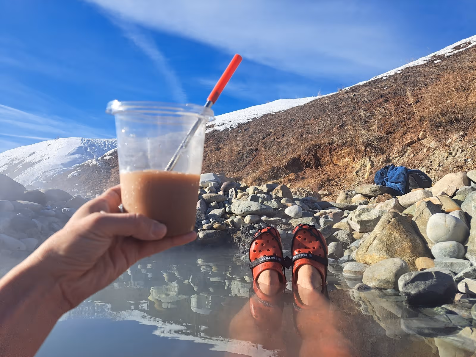 Steaming natural hot spring soaking pool surrounded by rocks with a hand holding a drink and feet wearing red shoes at Astoria Hot Springs.