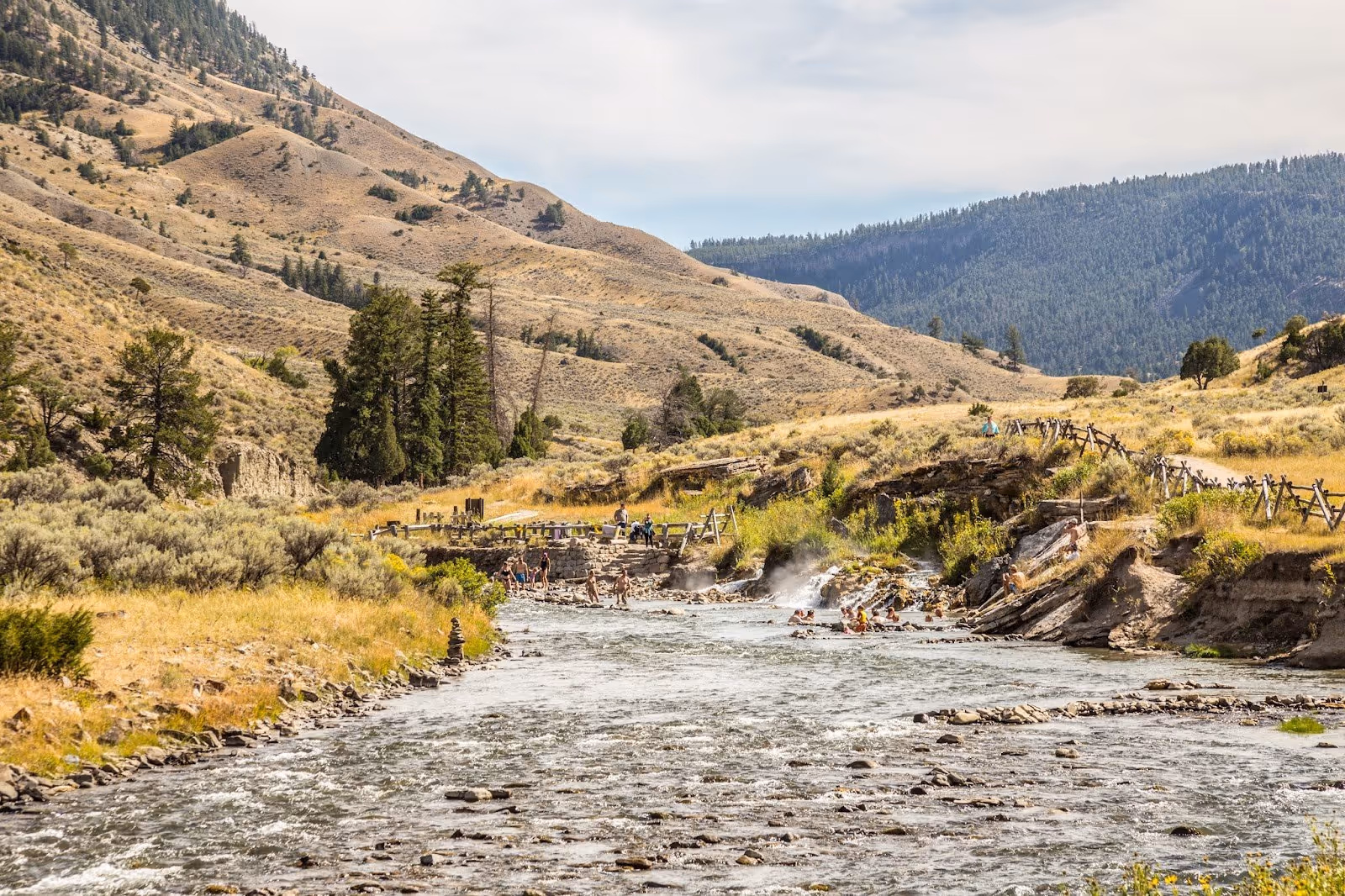 Wide view of Boiling River Hot Springs with groups of people soaking in pools along the flowing river surrounded by sagebrush and dry hills.