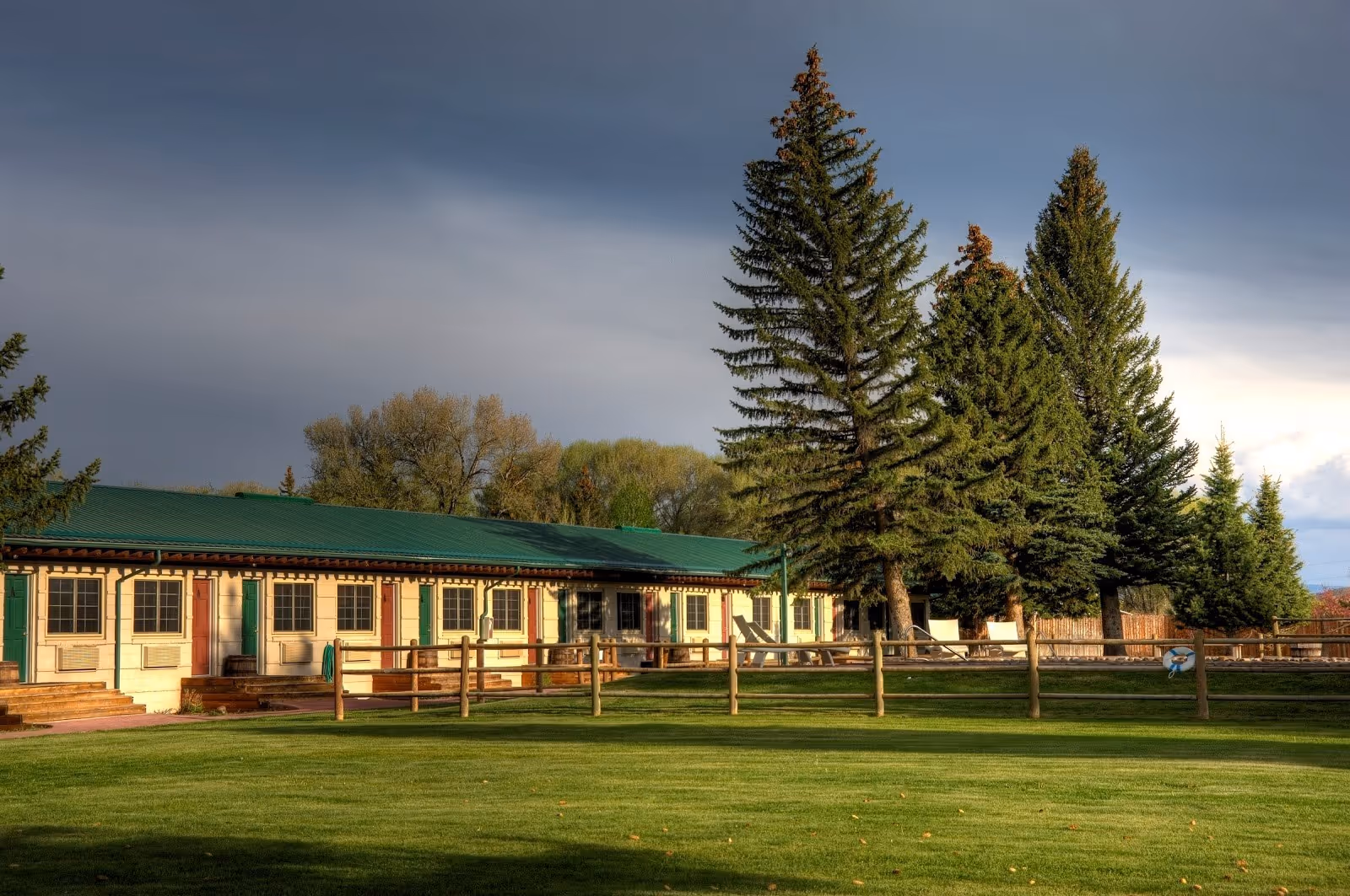 Long building with green roof and multiple doors facing a fenced grass area with tall pine trees at Saratoga Hot Springs Resort in Wyoming.