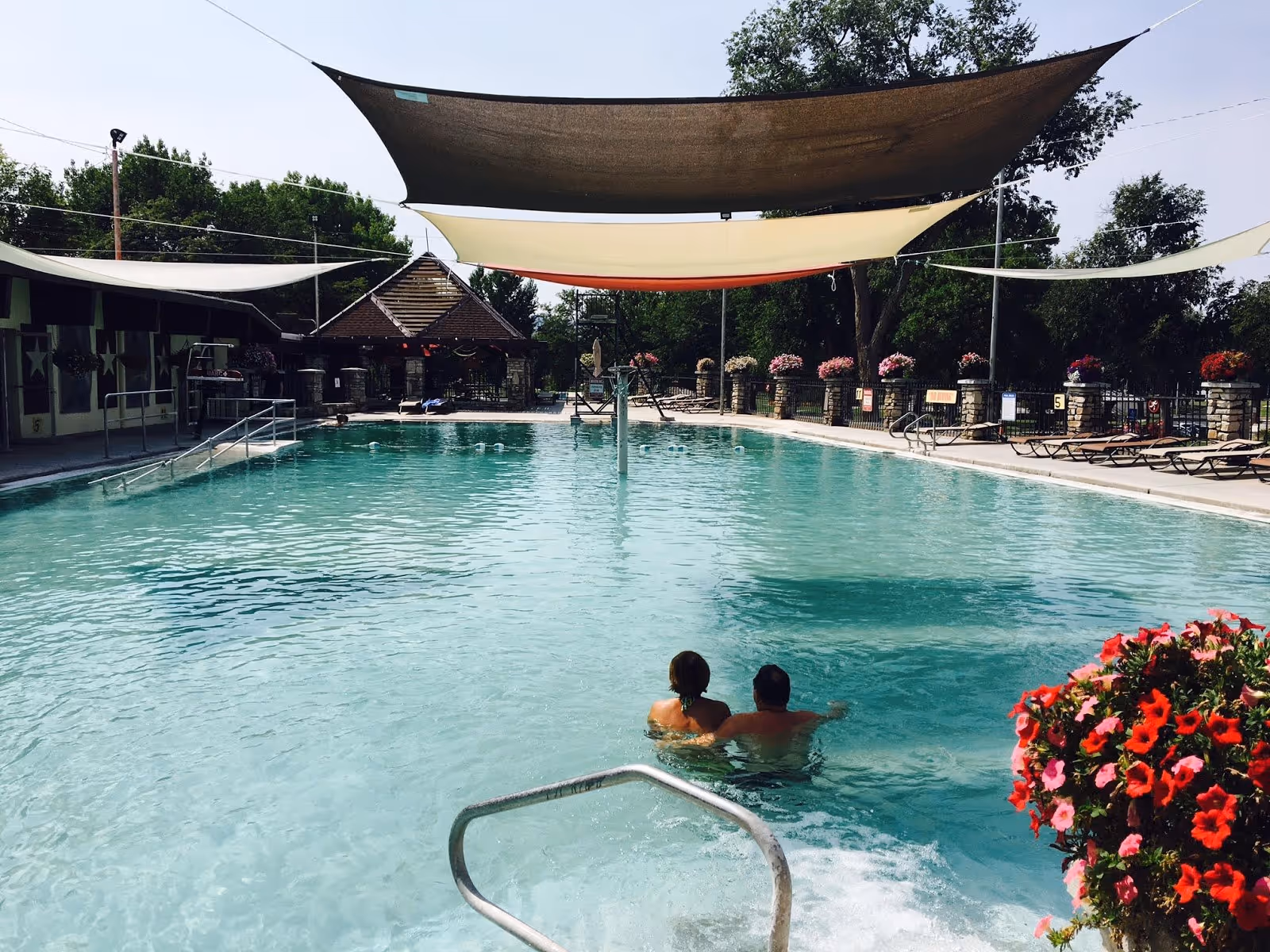 Couple soaking in the large outdoor hot spring pool at Star Plunge with shade sails and flowering plants in Thermopolis Wyoming.
