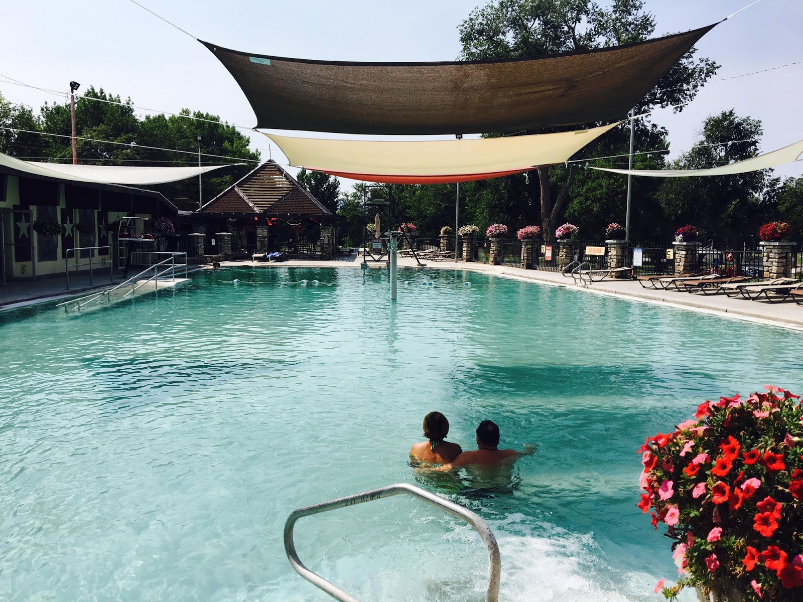 Couple soaking in the large outdoor hot spring pool at Star Plunge with shade sails and flowering plants in Thermopolis Wyoming.