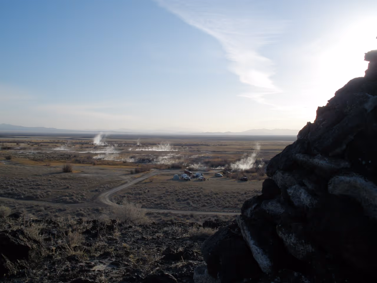 Wide view of Baker Hot Springs showing multiple steam vents rising over flat plains with mountain ranges in the faint distance.
