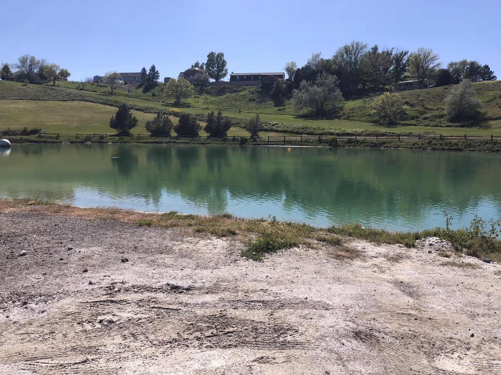 Calm mineral pool reflecting trees and houses on a hillside at Belmont Hot Springs RV Park near Garland, Utah under a clear sky.