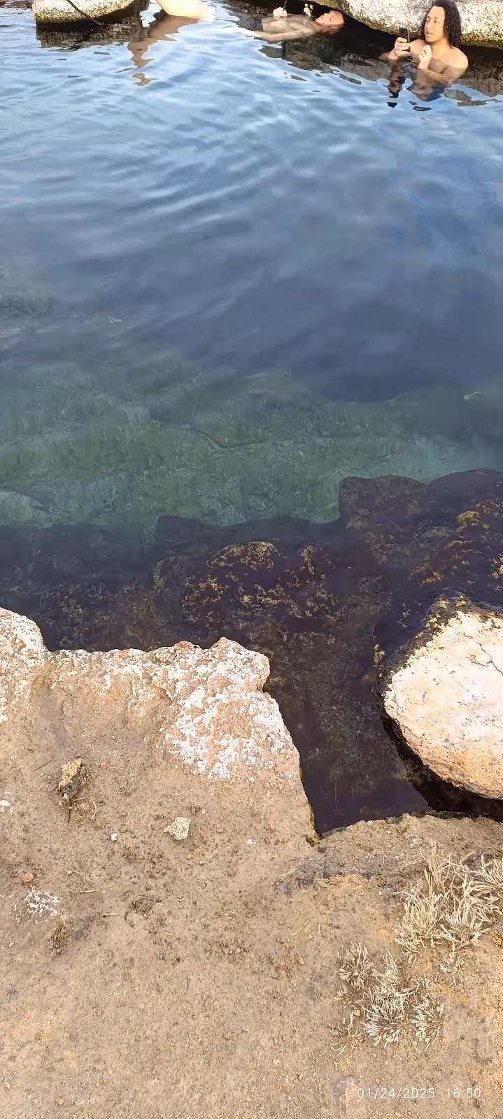 Soaking pool at Meadow Hot Springs with individuals relaxing near rocky ledges and clear mineral water visible below.