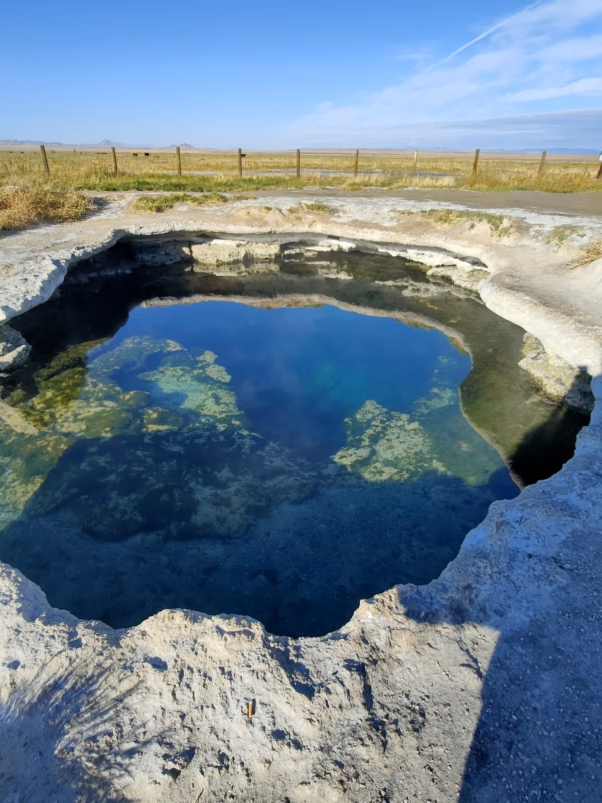 Natural hot spring pool with clear deep blue water surrounded by rocky edges and a wooden fence near Fillmore Utah.