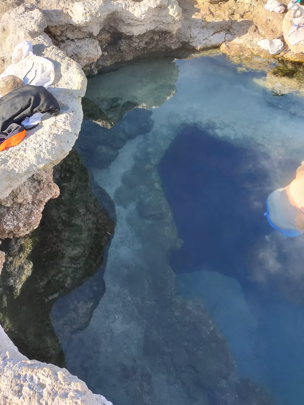 Rock-walled hot spring pool with people soaking, clothes resting on the edge, and clear water showing different depths.
