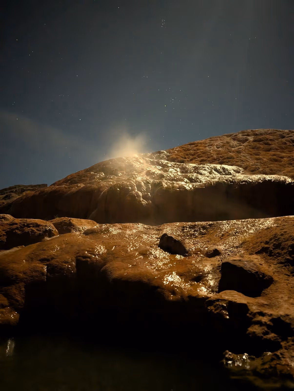 Steam rising from warm Red Hill Hot Springs mineral pools at night under a starry sky with light reflecting off rocky formations.