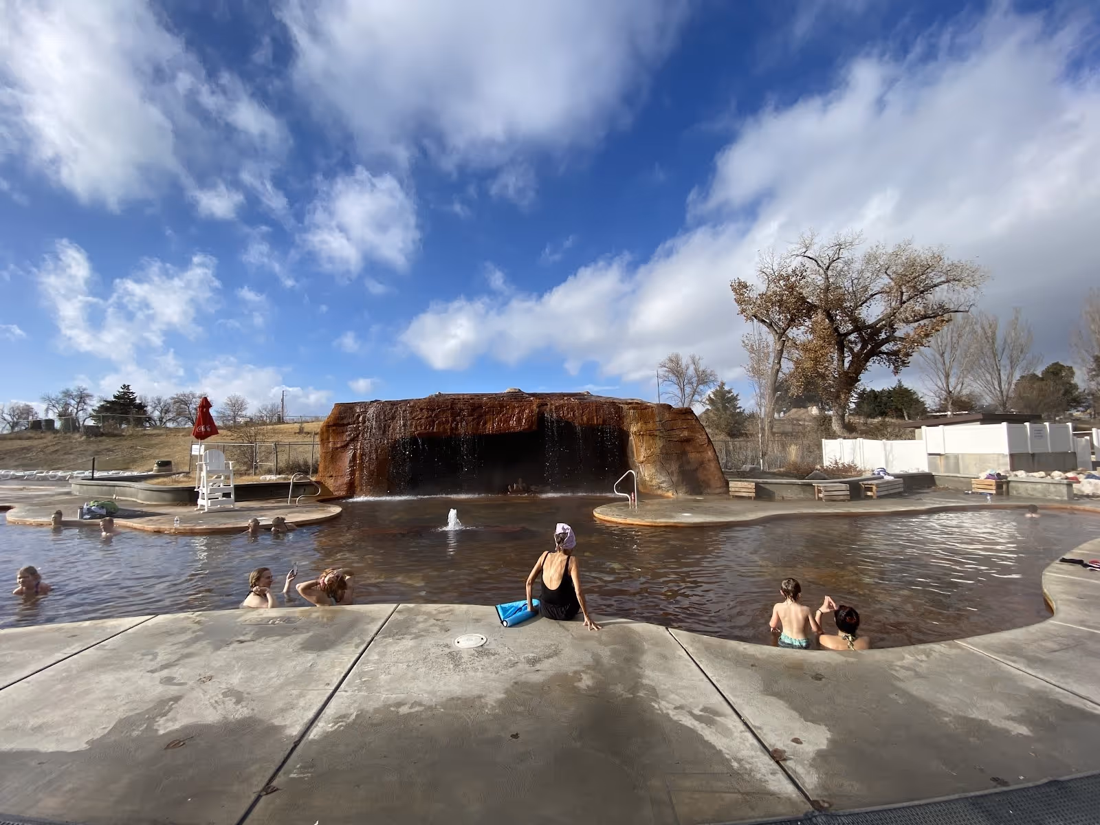 Outdoor Roosevelt Hot Springs pool with a rock waterfall feature and visitors soaking under a partly cloudy sky near Milford Utah.