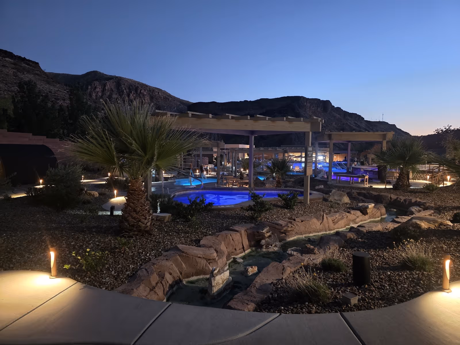 Night view of illuminated hot spring pools and dry landscaped garden beds with palm trees and distant mountains at Zion Canyon Hot Springs.