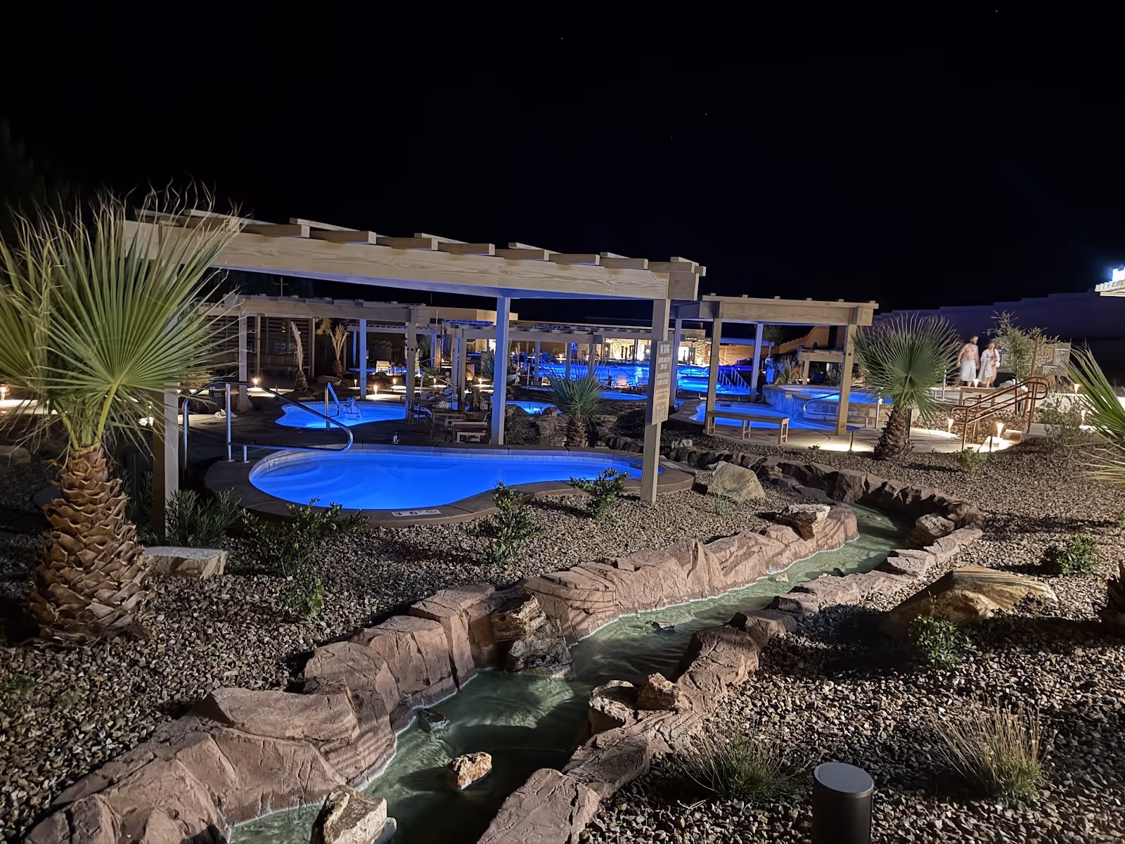 Blue-lit hot spring pools under wooden pergolas at night with desert plants and a rocky water channel at Zion Canyon Hot Springs Utah.