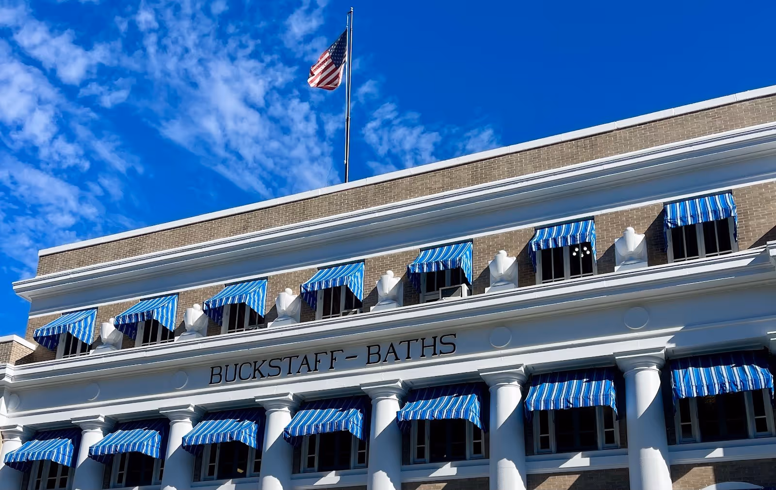 Buckstaff Bathhouse in Hot Springs Arkansas features white columns and blue striped awnings under a partly cloudy sky with an American flag.