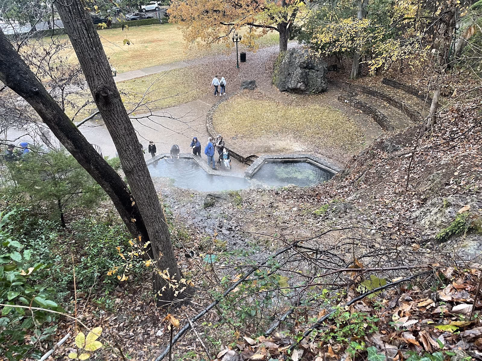 Overhead view of people gathered beside a steaming hot spring soaking pool with stone edges in a park setting near Hot Springs, Arkansas.