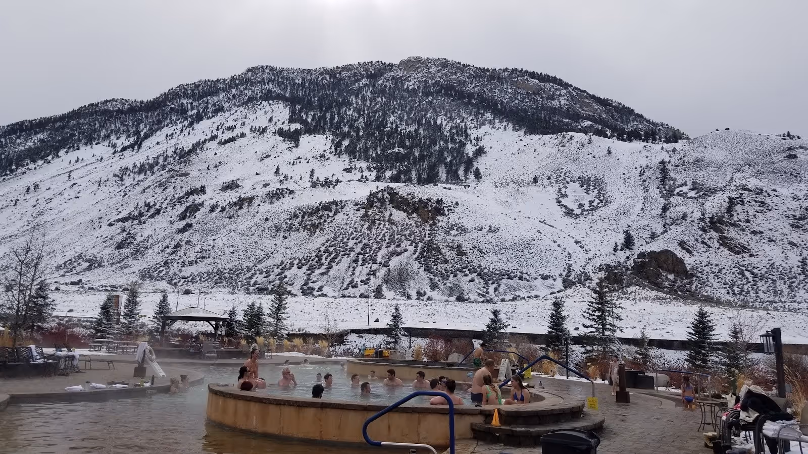 Steam rising from people soaking in circular hot spring pools with mountain landscape covered in light snow at Corwin Hot Springs Montana.