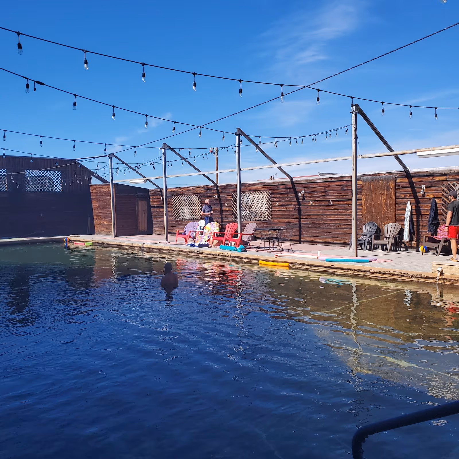 Sunny open-air hot spring pool with clear water, wooden deck, chairs, and string lights at Jackson Hot Springs Lodge in Montana.
