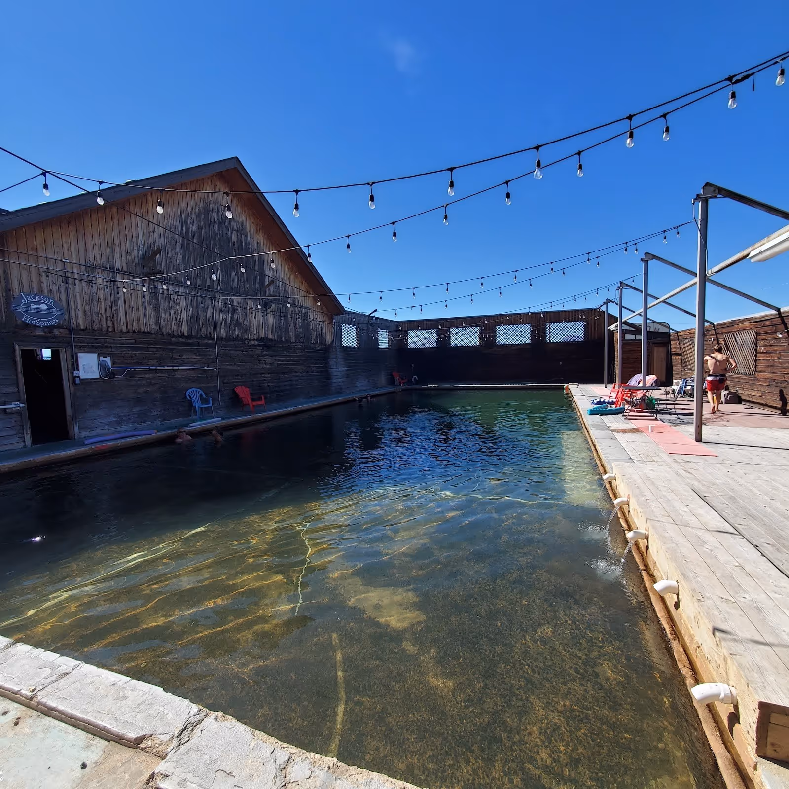 Steaming mineral pool with clear water and wooden deck seating under string lights at Jackson Hot Springs Lodge near Jackson Montana.