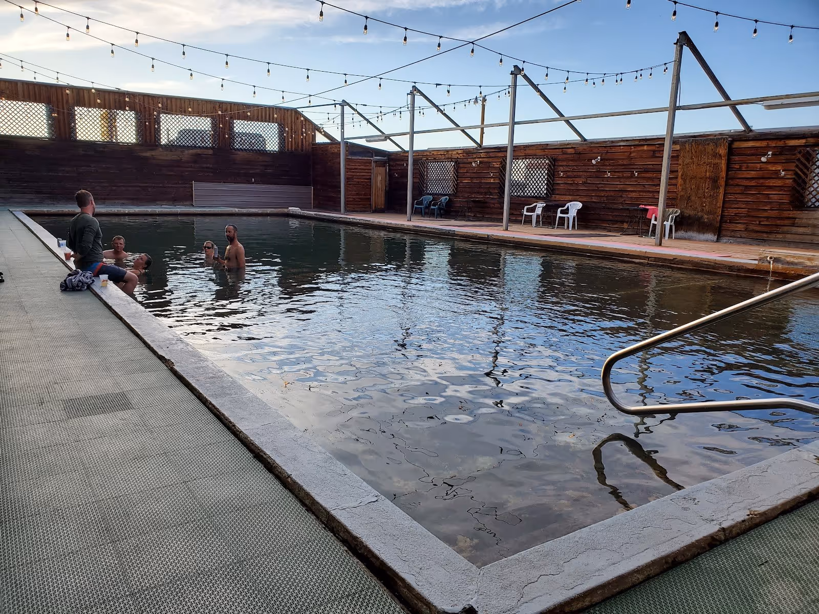 Guests soaking in a large rectangular hot spring pool with stairs, wooden deck, and overhead string lights at Jackson Hot Springs Lodge.