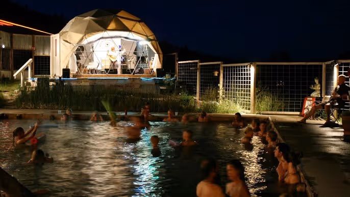 Night view of Norris Hot Springs with lit domed structure beside a crowded outdoor hot spring soaking pool in Norris Montana.