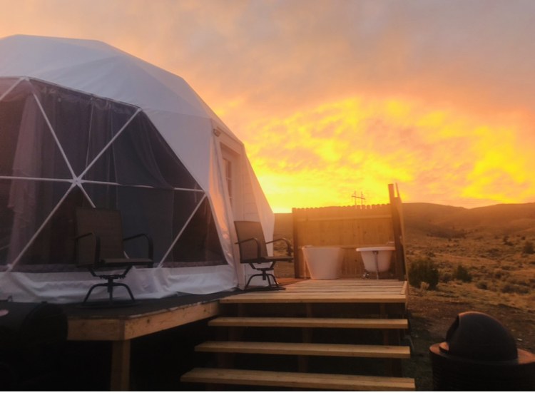 White soaking tubs on a raised wooden platform beside a dome tent under a colorful sunset at Pipestone Hot Springs near Whitehall Montana.