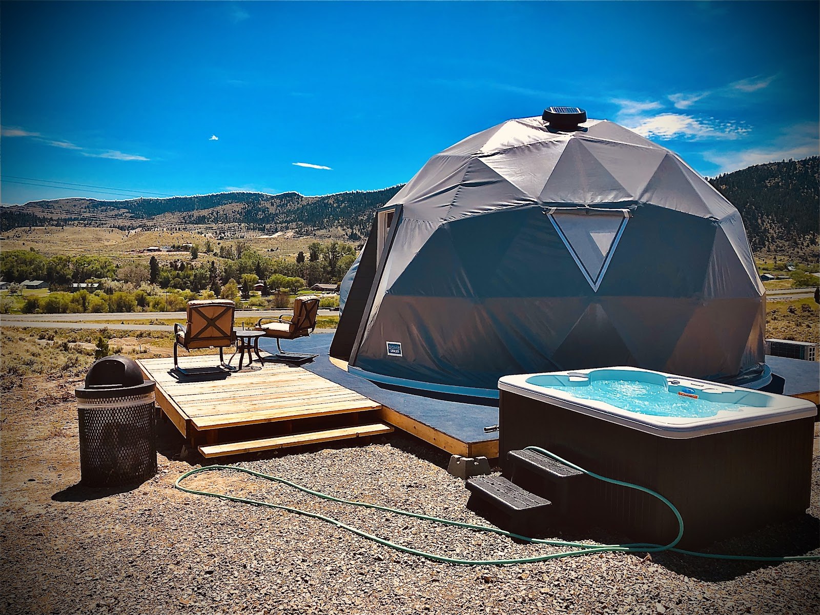 Outdoor hot tub next to a large geodesic dome with chairs on a wooden deck and montana mountains in the background at Pipestone Hot Springs.