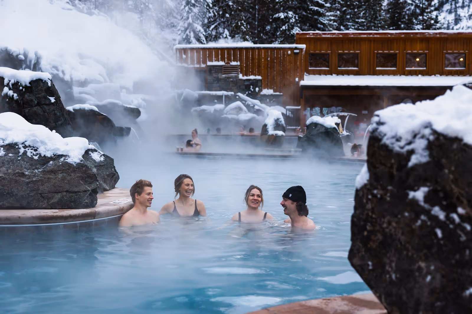 Four people relax in a steaming outdoor hot spring pool surrounded by snow-covered rocks at Quinn's Hot Springs Resort in St. Regis Montana.