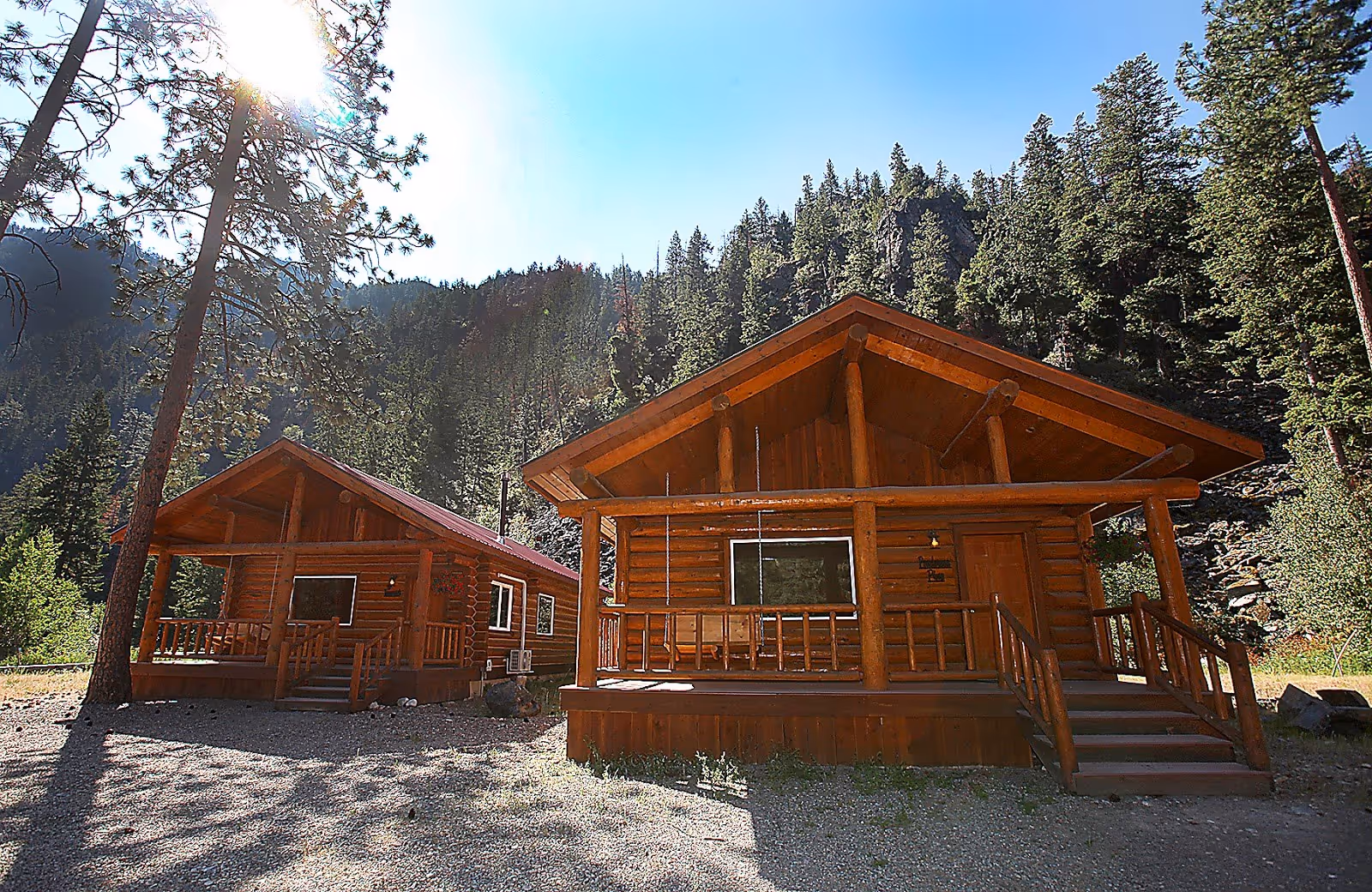 Two wooden log cabins with porches sit under bright sunlight near a forested mountain at Quinn's Hot Springs Resort, Montana.
