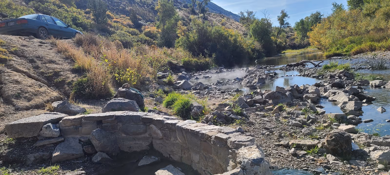 Stone-walled hot spring pool emitting steam next to a flowing river and green vegetation at Renova Hot Springs, Montana.