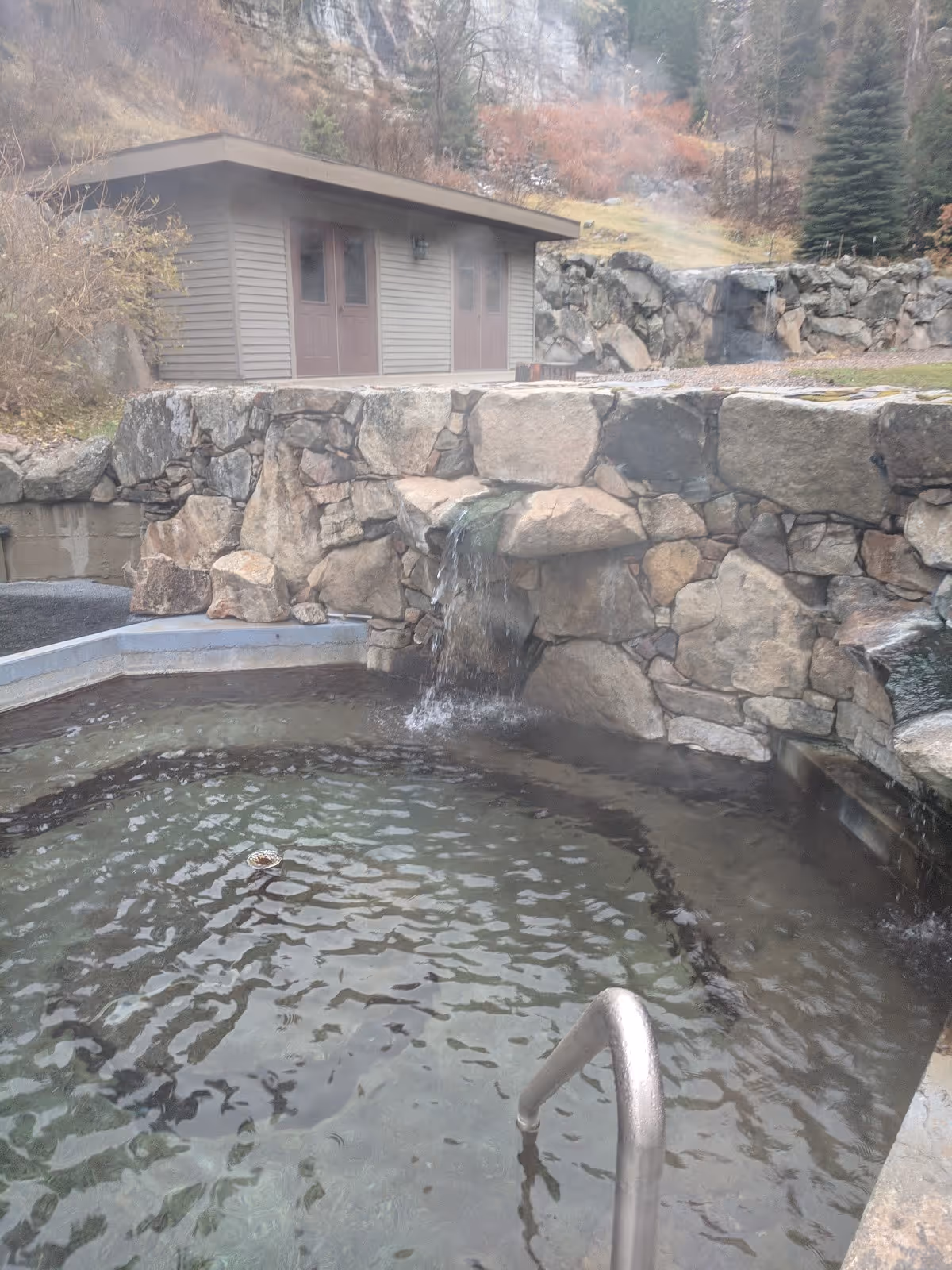 Small round soaking pool with stone wall waterfall feature and metal handrail steaming at Sleeping Child Hot Springs montana.