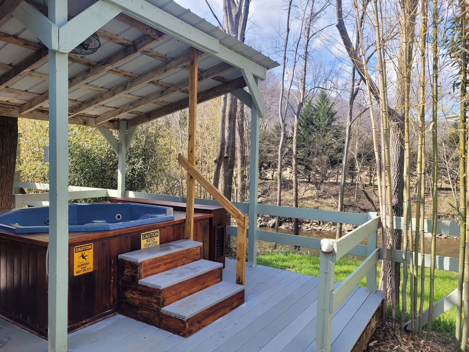 Octagonal blue spa pool set in a wooden structure next to a fire pit and black chair, with a view of a river and bare trees around Hot Spri.