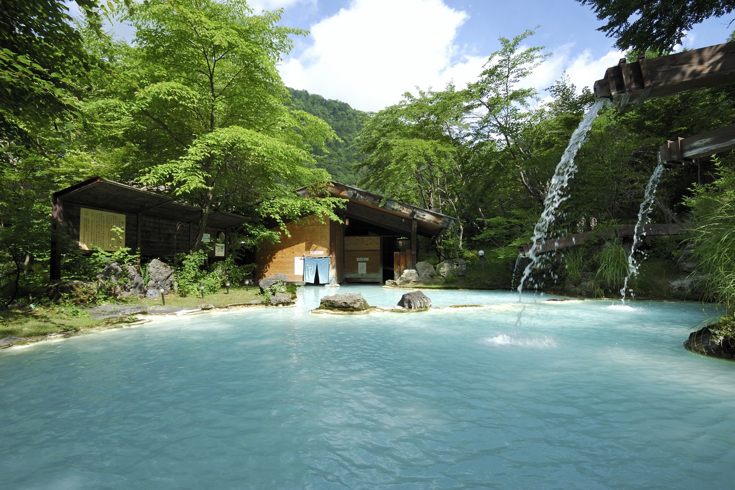 Awanoyu hot spring pool with water flowing from wooden spouts and lush green forest on a sunny day near Matsumoto, Japan.