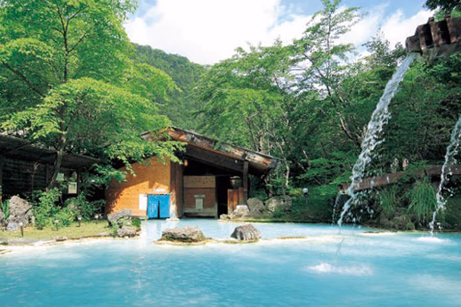Light blue mineral pool with small rocky islands and a wooden building backdrop at Awanoyu, near Matsumoto, Japan.