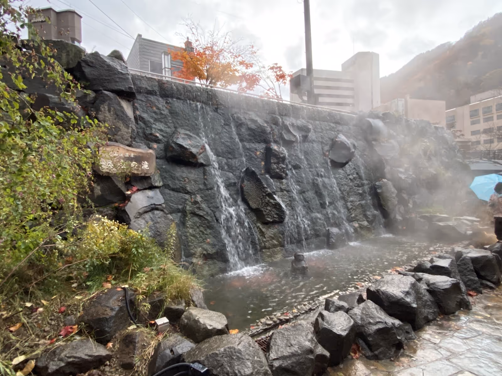 Steam rises from a natural hot spring waterfall cascading down a stone wall with surrounding foliage in Jozankei Onsen Japan.