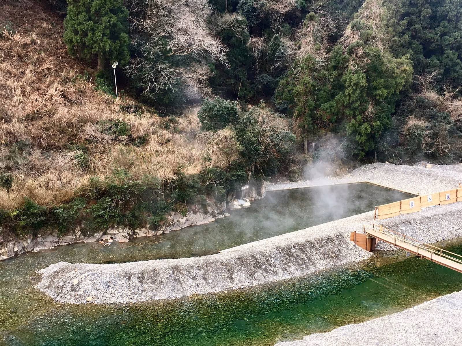 Steam rising from a natural hot spring river pool with green riverbed and forested hills near Tanabe, Japan.