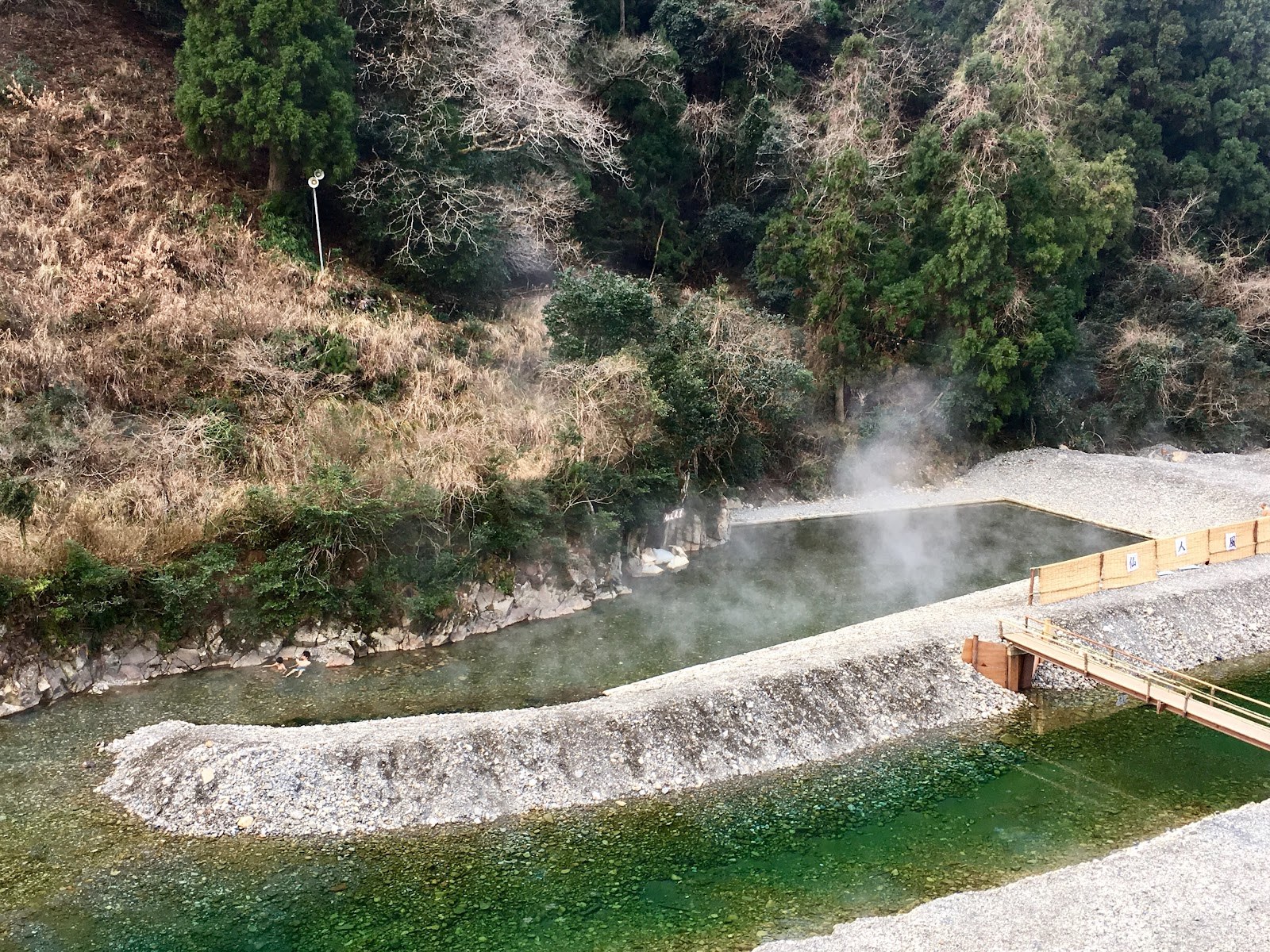 Steam rising from a natural hot spring river pool with green riverbed and forested hills near Tanabe, Japan.