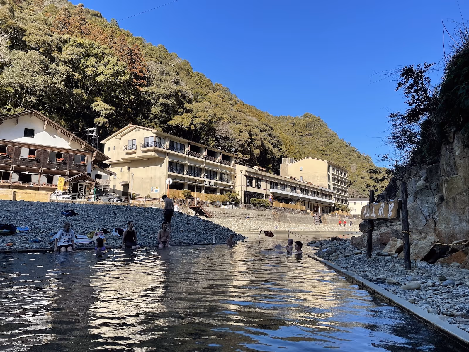 Visitors bathing in Kawayu Onsen Sennin-buro River Bath along a rocky river surrounded by buildings in Tanabe, Japan.