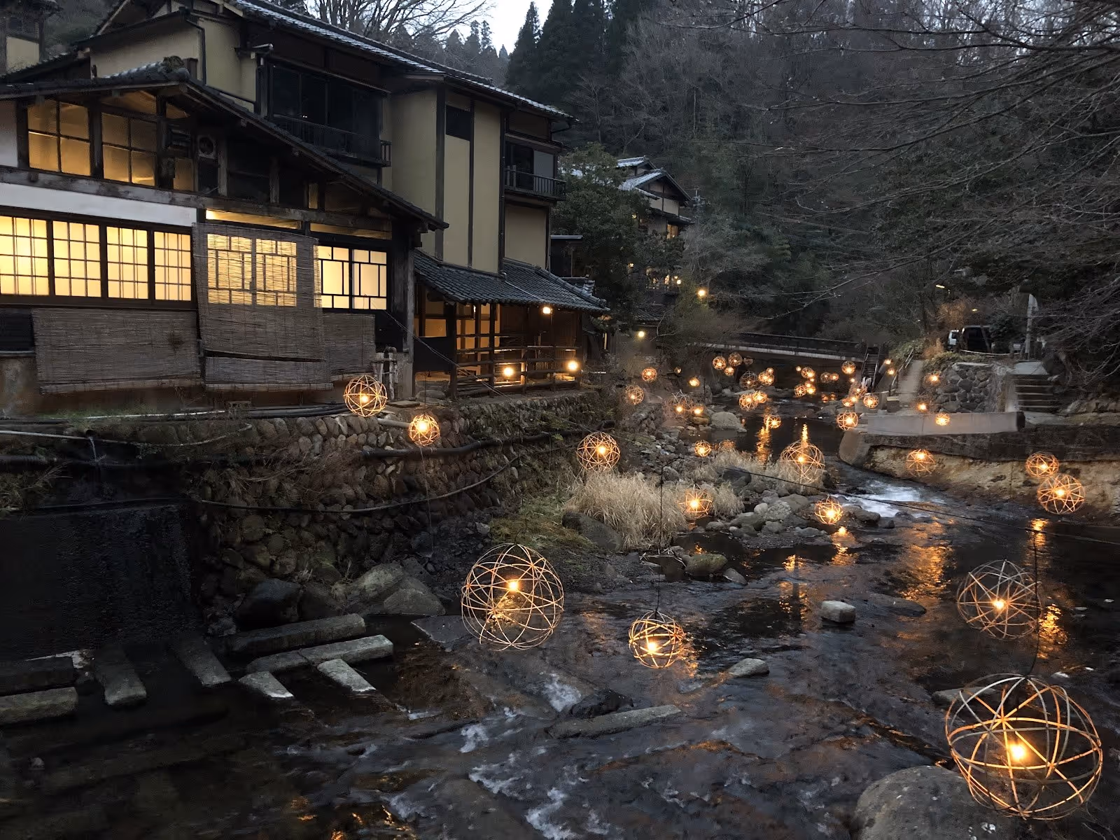 Evening view of a river near traditional buildings decorated with glowing round lights in Kurokawa Onsen, Japan.