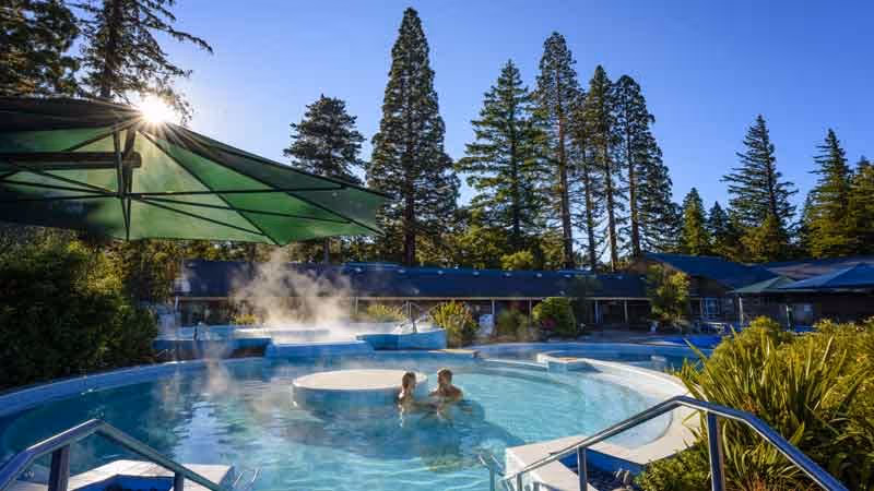 A close-up view of people enjoying steaming hot spring pools with greenery and large umbrella shade at Hanmer Springs near Hanmer Springs N.