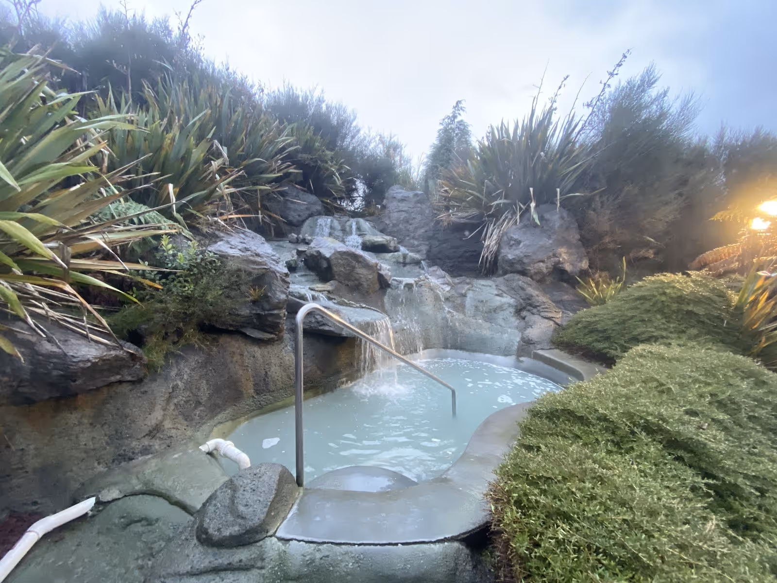 Heated spa pool with a cascading waterfall surrounded by lush green bushes and large rocks at Hell's Gate Mud Bath & Spas.