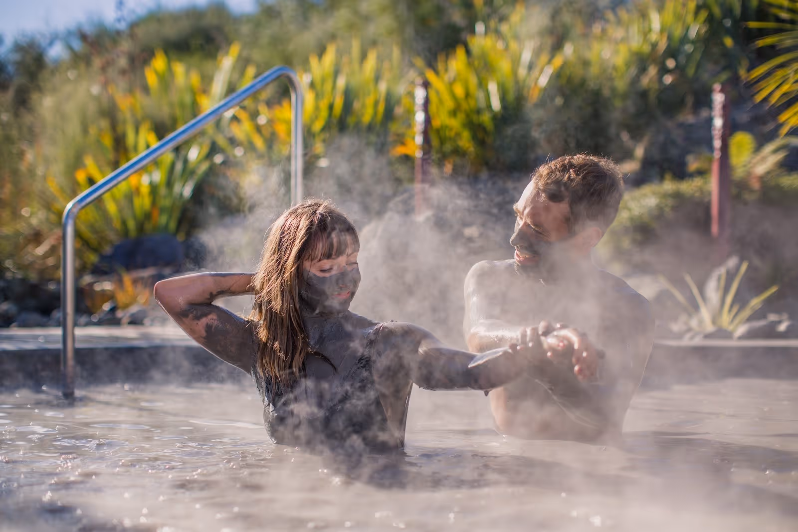 A man and woman covered in mud relax in a steaming hot spring pool at Hell's Gate Mud Bath & Spas in Rotorua New Zealand.