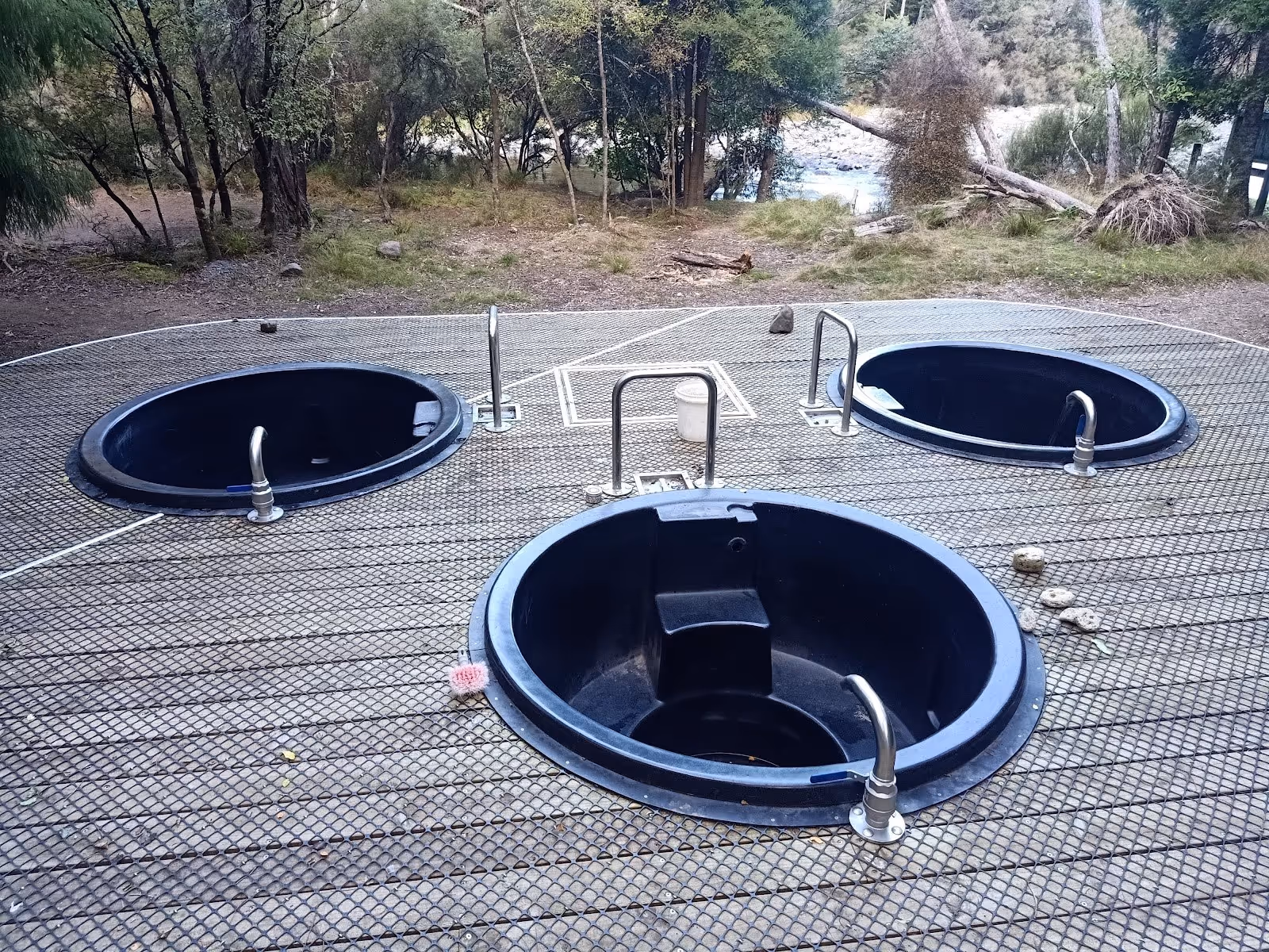 Three circular black hot spring pools set on a textured metal platform with handrails, located outdoors near forest in New Zealand.