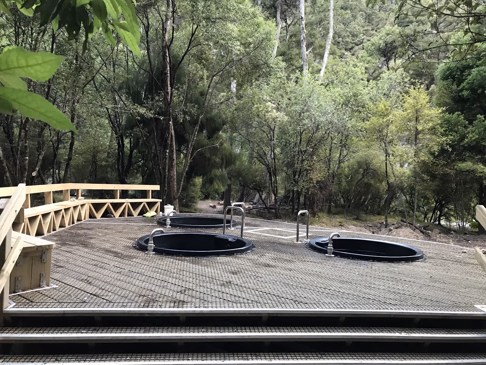 Three round black soaking pools on a mesh platform next to steps with green trees in the background at Mangatainoka Hot Springs.