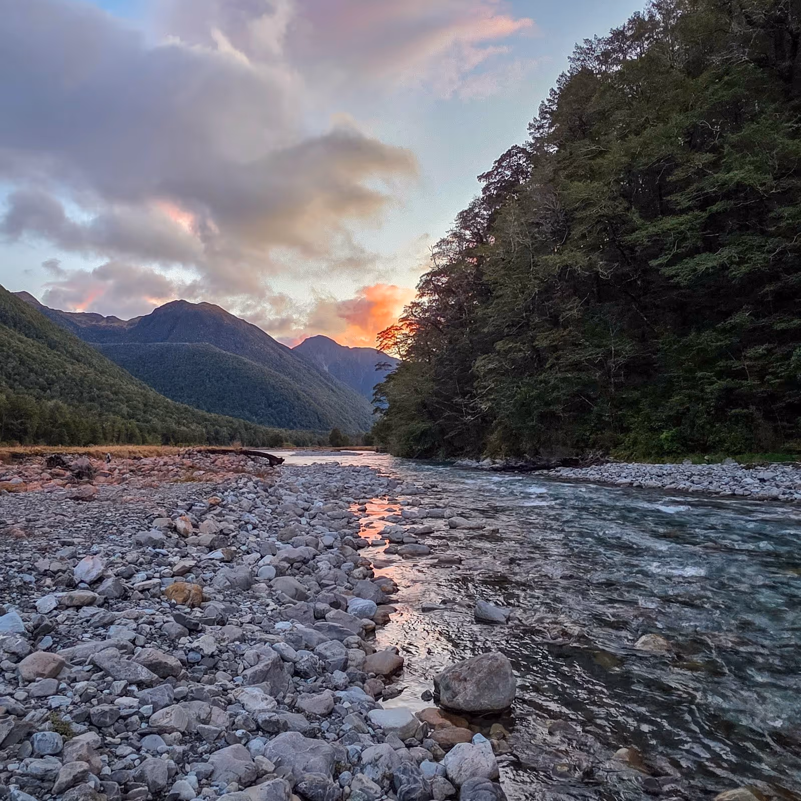 Rocky riverbank at sunset with flowing water and forested hills at Maruia Hot Springs area near Springs Junction New Zealand.