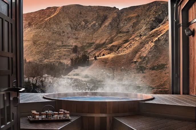 Steam rising from a circular wooden hot spring pool at Onsen Hot Pools overlooking rugged mountains near Queenstown.