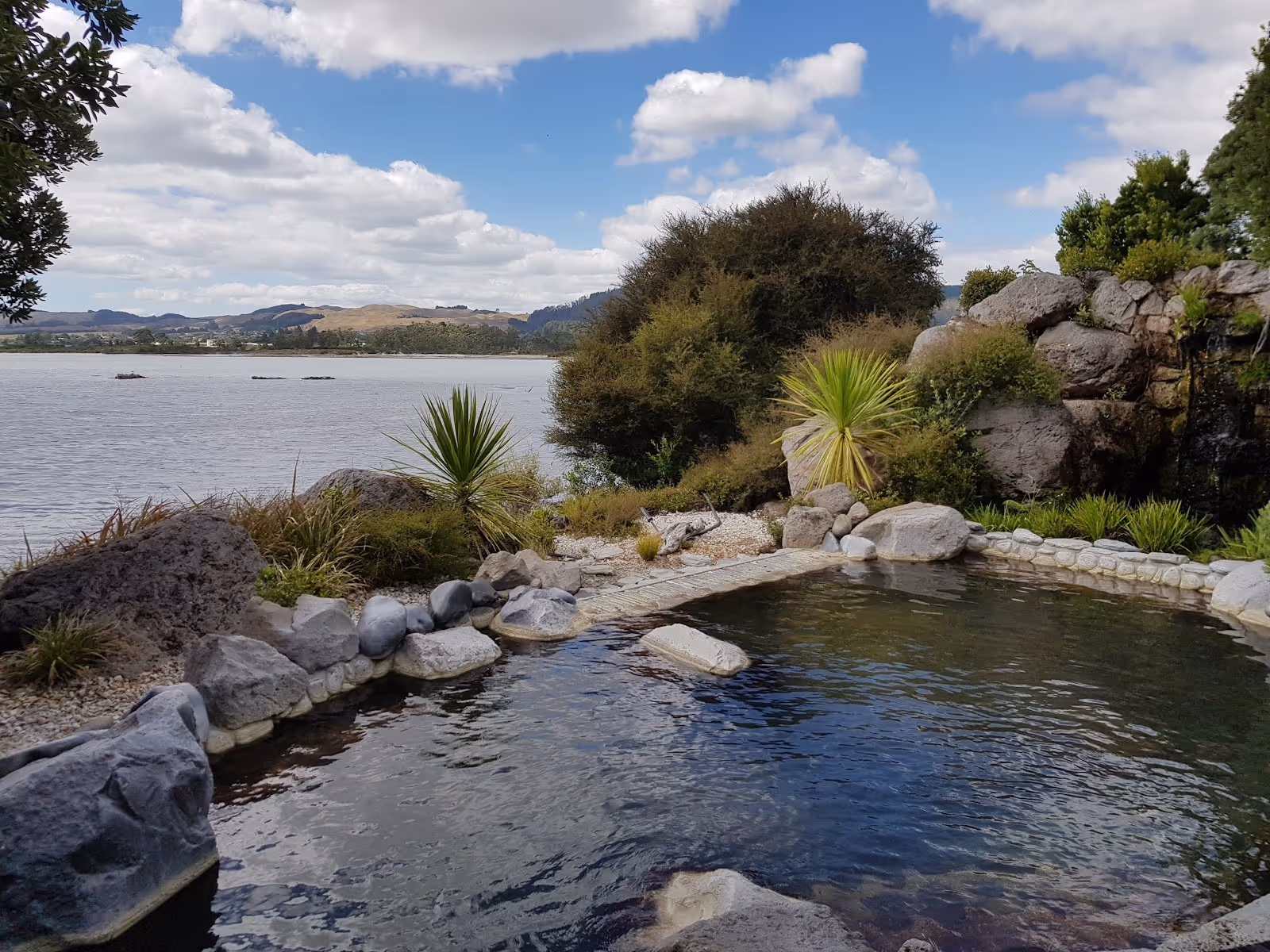 Natural-looking outdoor hot spring pool bordered by stones near lake shore with bushes and hills in the Rotorua area of New Zealand.