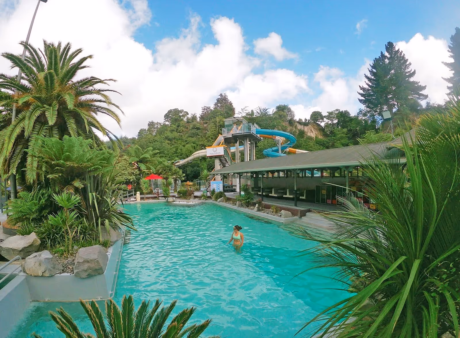 Large turquoise hot spring pool with water slide tower and lush greenery around at Taupo DeBretts Hot Springs near Taupō New Zealand.