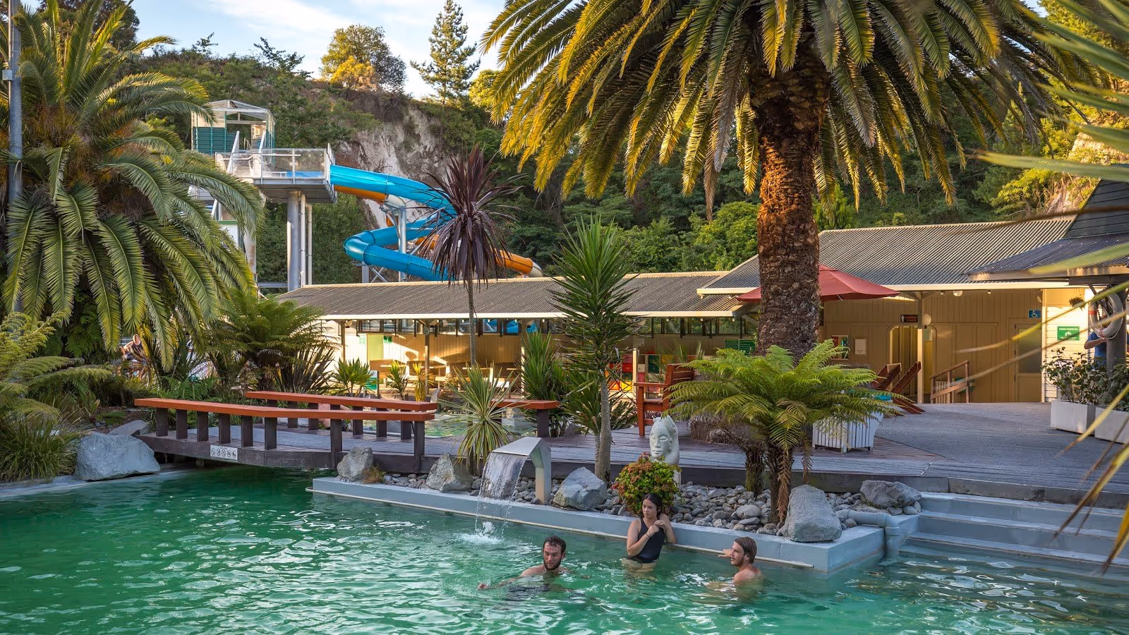 Outdoor hot spring pool with green water surrounded by palm trees and a water slide in Taupō New Zealand at Taupo DeBretts Hot Springs.