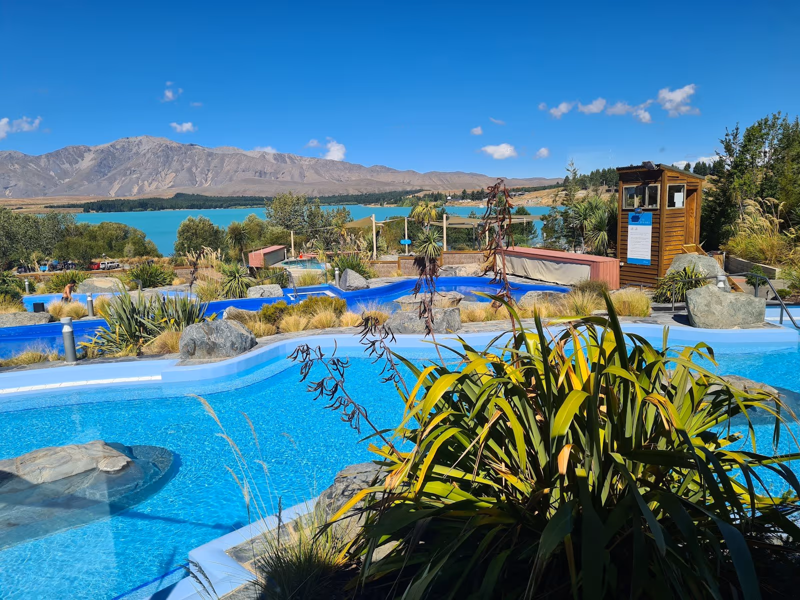Curved pools at Tekapo Springs offer views of Lake Tekapo and distant mountains with surrounding plants and rock features in New Zealand.