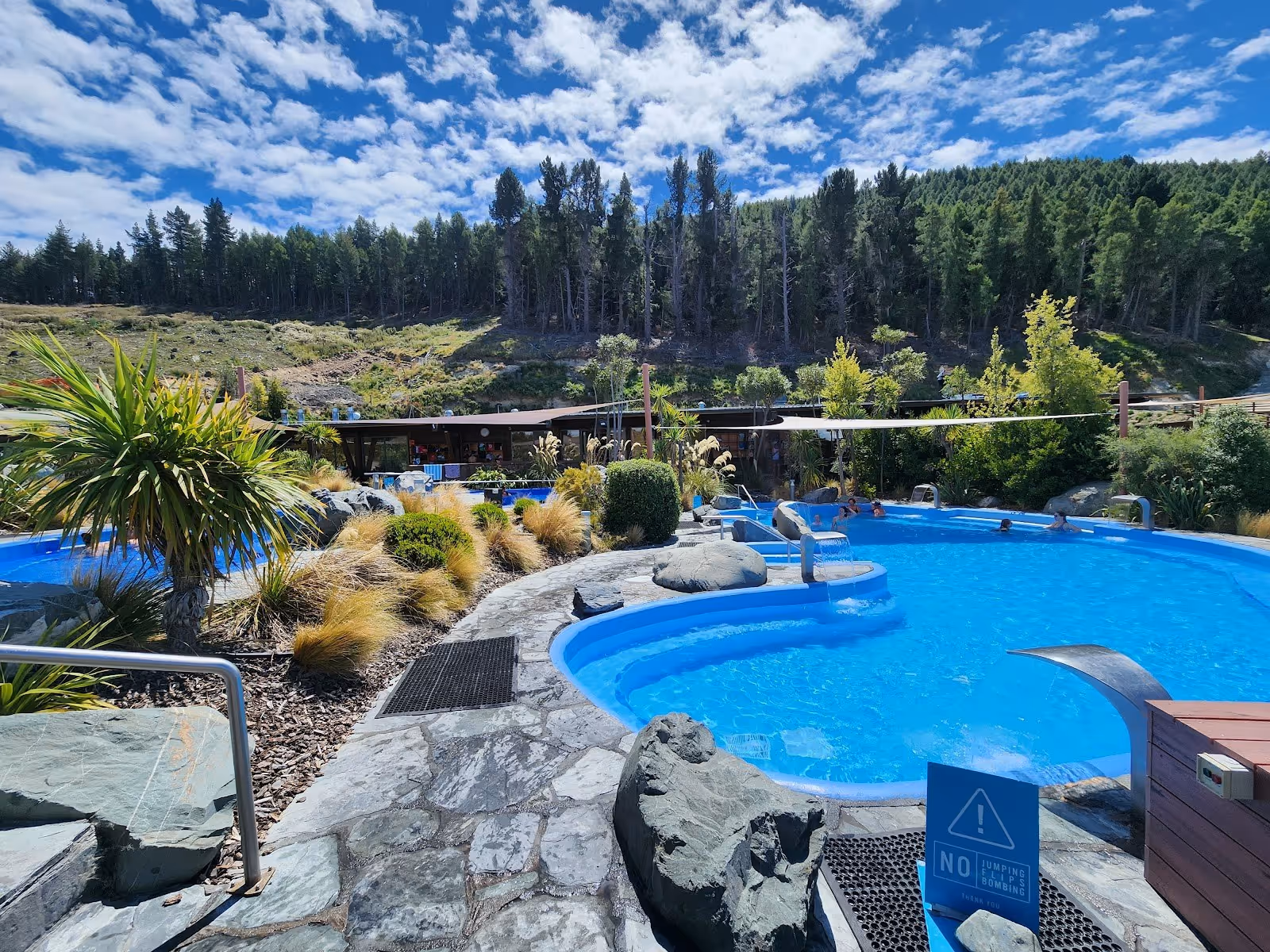 Bright blue mineral pools at Tekapo Springs are set against a backdrop of dense green hills and a partly cloudy sky near Lake Tekapo New Ze.